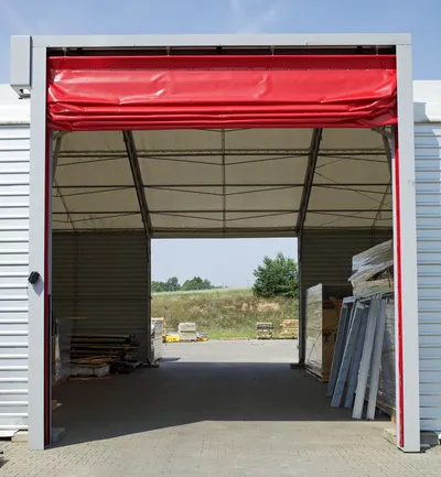 La imagen muestra una puerta enrollable de lona roja en un entorno industrial. Este tipo de puertas ofrecen una solución rápida y eficiente para el acceso en áreas de alto tráfico, manteniendo la seguridad y el control climático.